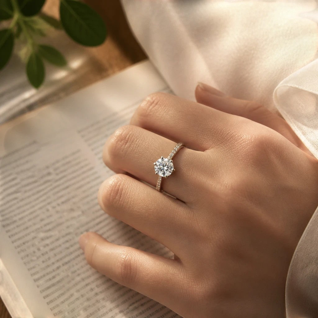 My Store solitaire engagement ring with a brilliant center diamond and pavé band on a woman's finger over a book.